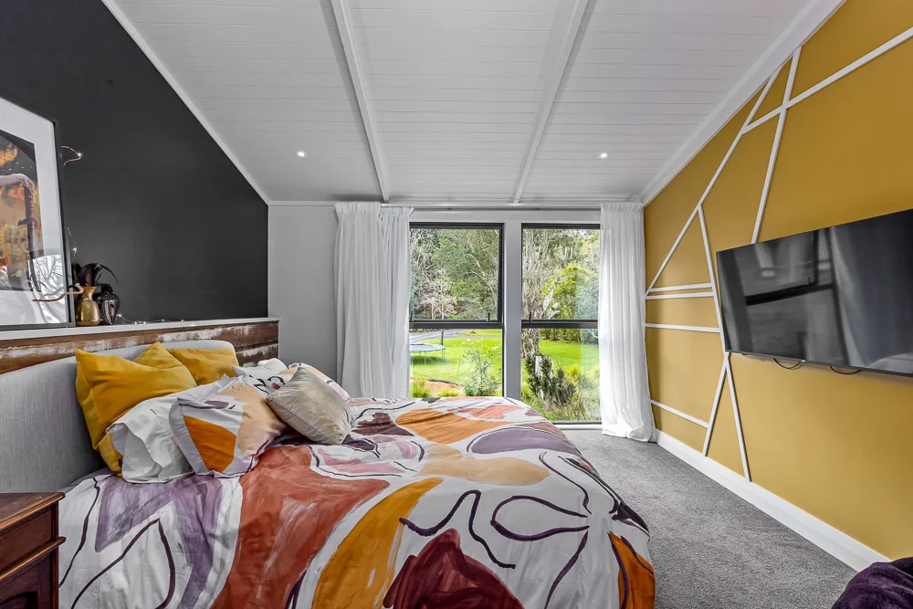 Auckland bedroom with mustard yellow geometric feature wall, tongue and groove ceiling and full-height window looking over the lawn