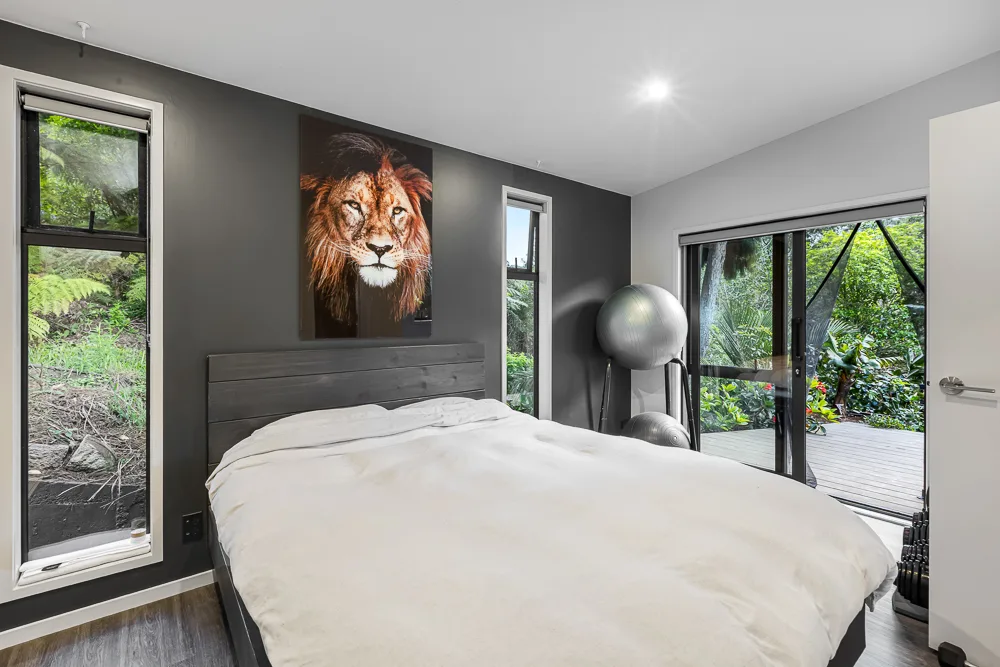Auckland bedroom with dark charcoal feature wall, tall narrow windows to native bush and sliding doors opening onto a timber deck