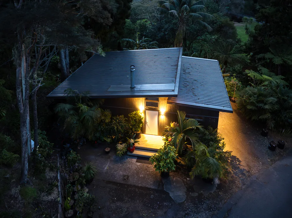 Elevated dusk view of an Auckland studio building with corrugated hip roof, lit entry and surrounding native bush